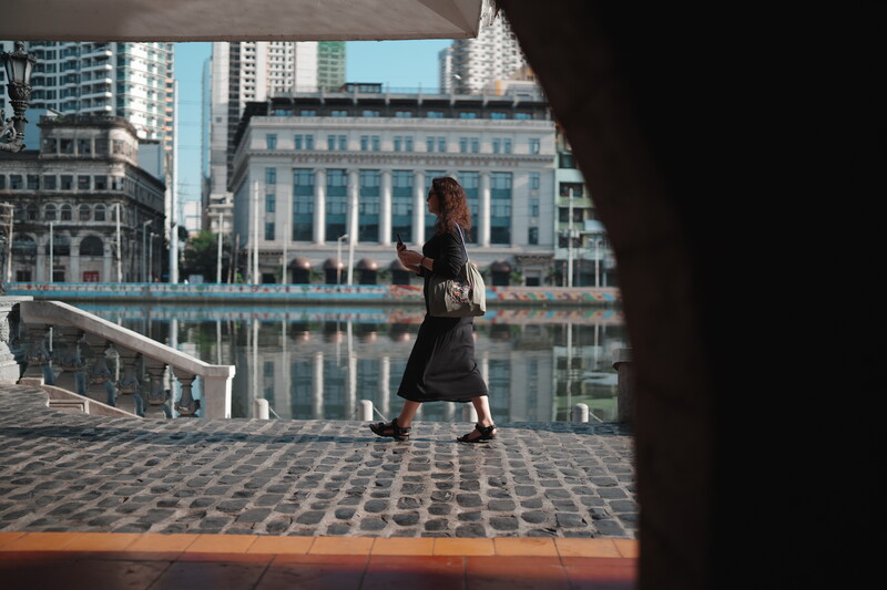 Woman walking on waterfront promenade framed by architectural foreground, urban skyline beyond