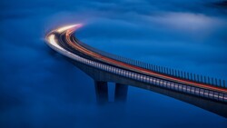 Long-exposure photograph of a curved bridge at dusk with vehicle light trails creating glowing arcs against a deep blue sky