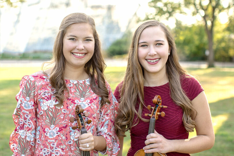 Two young musicians holding violins outdoors in a park setting